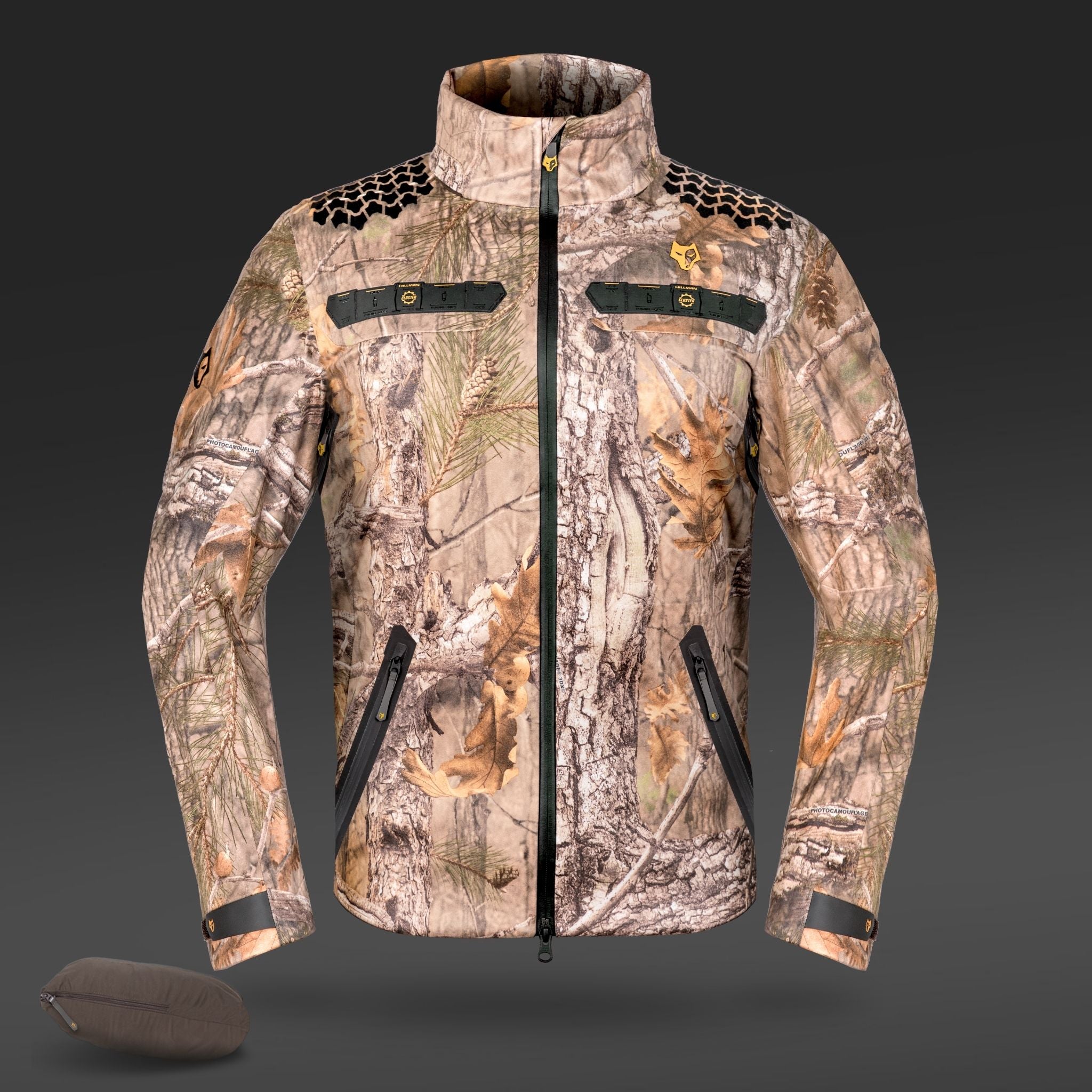 Die Hillman 5WL Ultraleichte Regenjacke besticht durch ein Camouflage-Muster mit Waldmotiven, Baumrinde und Blättern. Sie verfügt über schwarze Reißverschlüsse, ein kleines rundes Logo am linken Arm und eine passende Tasche zur einfachen Aufbewahrung.