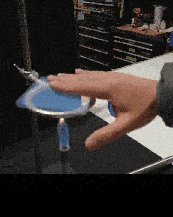 Eine Person versucht, in einer Werkstatt einen blauen Stuhl auf einer Metallstange zu balancieren. Der Stuhl wackelt und bleibt kurz im Gleichgewicht, bevor er fast herunterfällt. Im Hintergrund sind Werkzeuge und Geräte in einem Lagerbereich zu sehen.