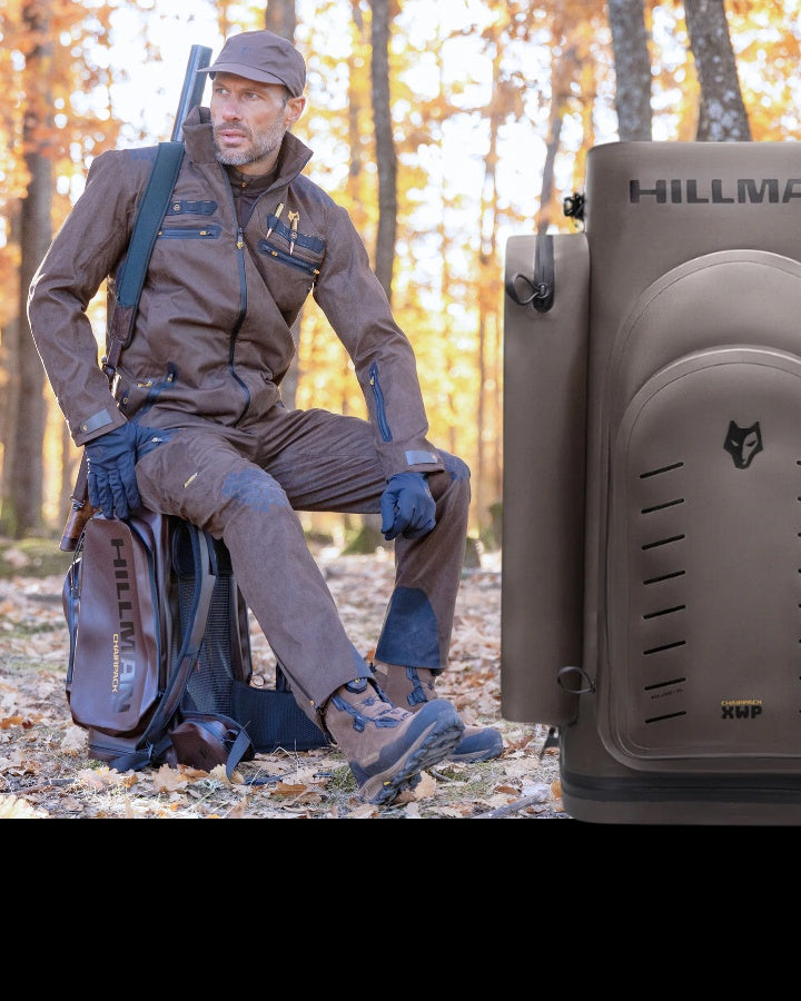 Mann im Freien im Herbstwald, trägt braun-blaue Outdoor-Ausrüstung und Mütze, hält Gewehr und Rucksack. Er kniet auf einem Knie auf gefallenen Blättern und blickt in die Ferne. Im Vordergrund ist ein Hartschalenkoffer mit der Marke „Hillman“ zu sehen.
