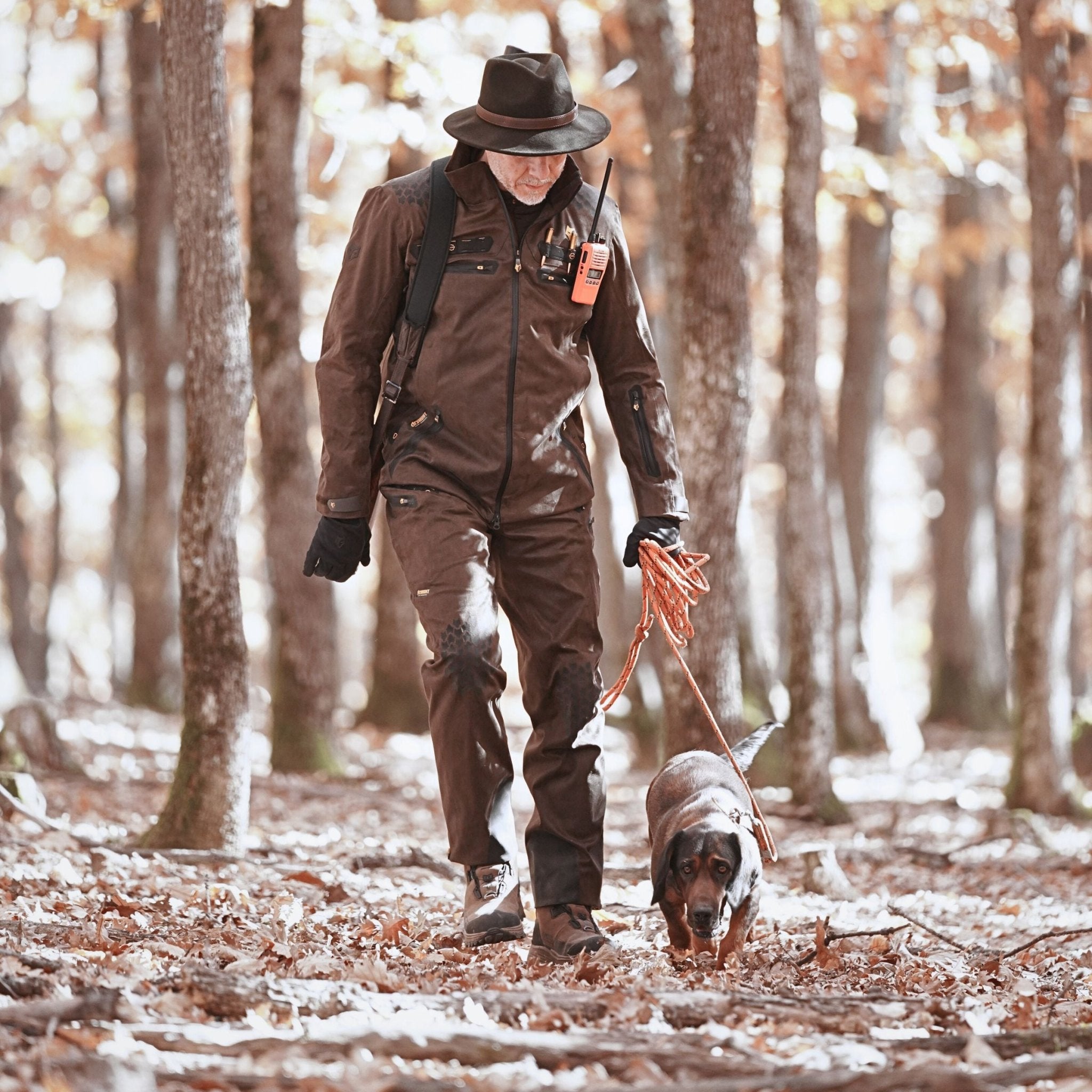 In einer Hillman 6OL Winter Jagdhose Camouflage führt eine Person einen schwarz-braunen Hund an der Leine durch einen laubbedeckten Wald. In der wasserdichten Jagdhose trägt sie einen Rucksack und ein Funkgerät, umgeben von hohen Bäumen im gesprenkelten Sonnenlicht.