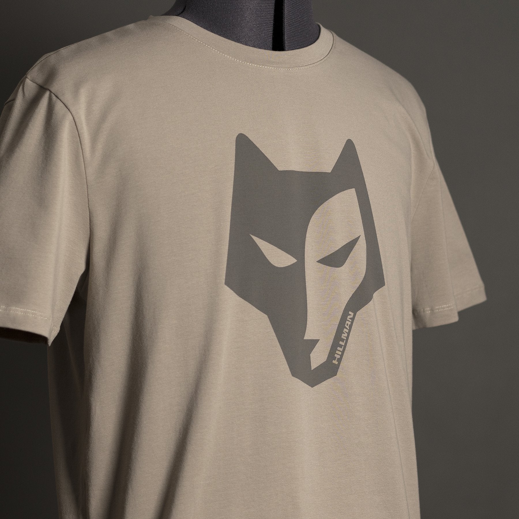 Das Hillman Jagdshirt Alpha - Jagd T-Shirt aus ägyptischer Baumwolle besticht durch eine stilisierte dunkelgraue Wolfsgrafik auf beigefarbenem T-Shirt. Präsentiert auf einer Schaufensterpuppe vor schlichtem dunkelgrünem Hintergrund, vereint es mit seinem kantigen Design Stil und Komfort perfekt.