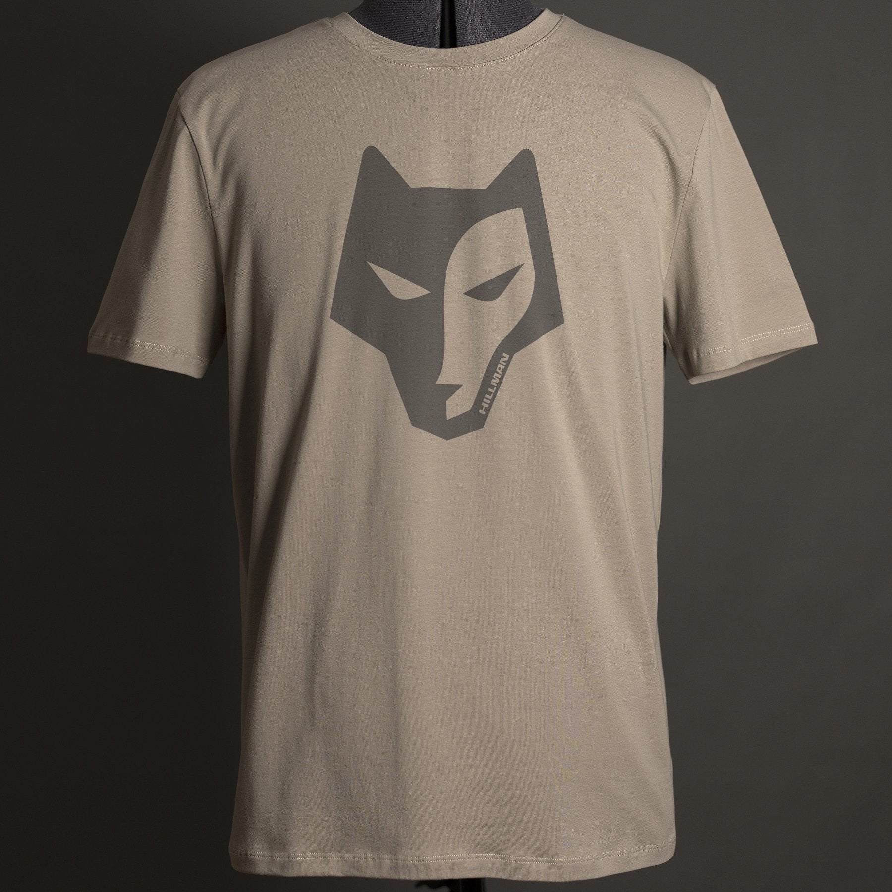 Das Hillman Jagdshirt Alpha – Jagd T-Shirt in Beige zeigt in der Mitte ein stilisiertes dunkles Wolfsgesicht. Es ist aus ägyptischer Baumwolle gefertigt und auf einer Schaufensterpuppe mit dunkelgrünem Hintergrund abgebildet, was die Hightech-Produktionsqualität unterstreicht.