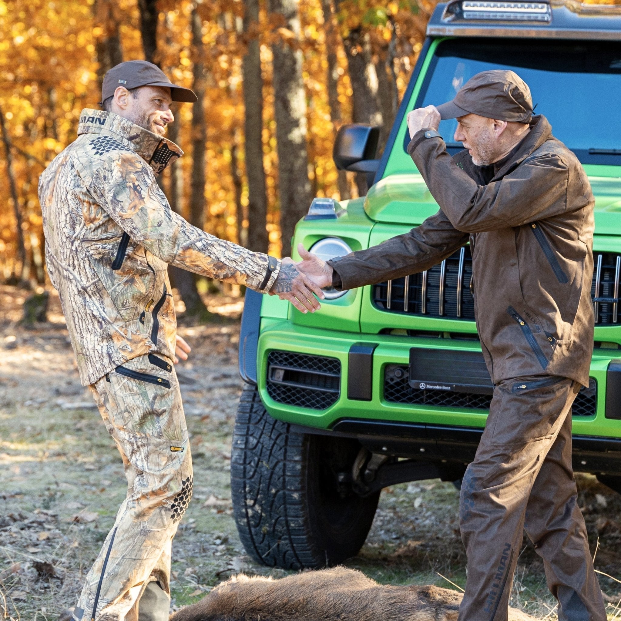 Zwei Jäger in funktioneller Hillman Jagdbekleidung begrüßen sich nach einer erfolgreichen Schwarzwildjagd im Wald vor einem grünen Geländewagen.