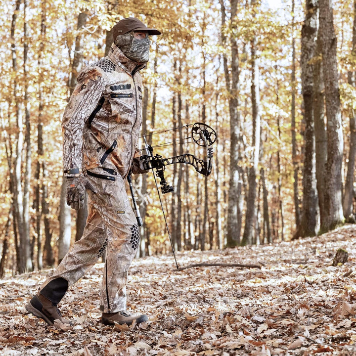 Eine Person in Tarnkleidung und mit Gesichtsbedeckung läuft mit einem Compoundbogen durch einen Wald, umgeben von Bäumen, deren Herbstlaub den Boden bedeckt.