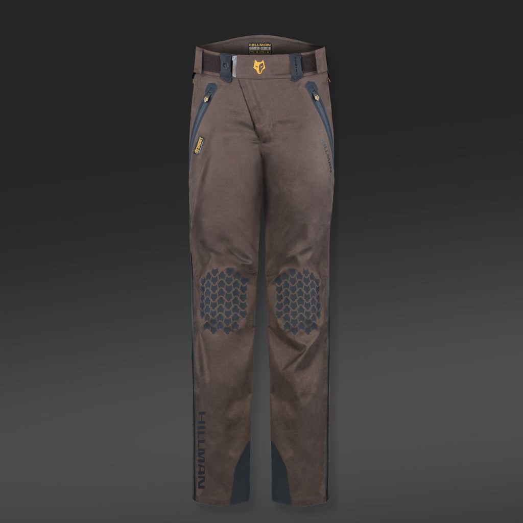 Die 5WL Wasserdichte Regenhose von Hillman ist eine ultraleichte braune Hose mit verstärkten Kniebesätzen, blauen Akzenten, Gürtelschlaufen, Reißverschlusstaschen und Jagdhosen-Branding auf dem Oberschenkel vor dunklem Hintergrund.