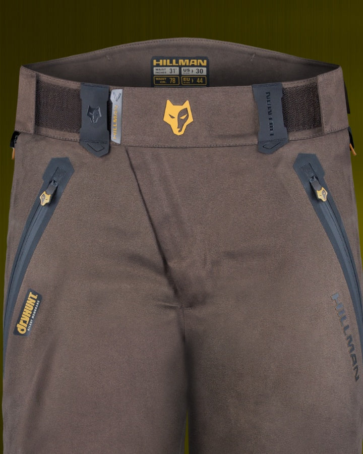 Braune Outdoorhose mit Reißverschlusstaschen und verstellbaren Gürtelschlaufen. Auf der Vorderseite befindet sich ein kleines gelbes Fuchslogo und auf den Taschen das „Hillman“-Branding. Der Hintergrund ist in einem gedeckten Grün gehalten.