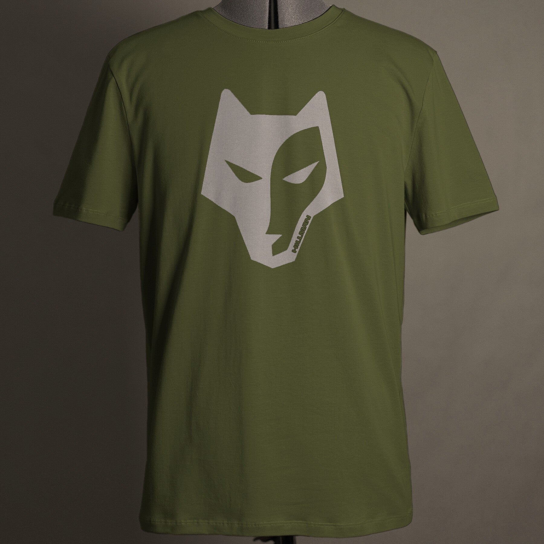 Das olivgrüne Jagdshirt Alpha von Hillman präsentiert sich mit einem stilisierten grauen Wolfsgesicht-Logo. Gefertigt aus hochwertiger ägyptischer Baumwolle vereint dieses T-Shirt Qualität und Stil. Präsentiert auf einer Schaufensterpuppe vor neutralem Hintergrund, verkörpert es mühelos Eleganz und Funktionalität.