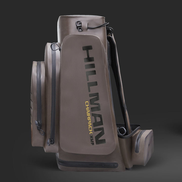 Abgebildet ist ein brauner Rucksack mit mehreren Fächern. Auf der Seite steht der Text „HILLMAN CHAIRPACK XHP“. Vor einem dunkelgrauen Hintergrund wirkt das Design robust und funktional.