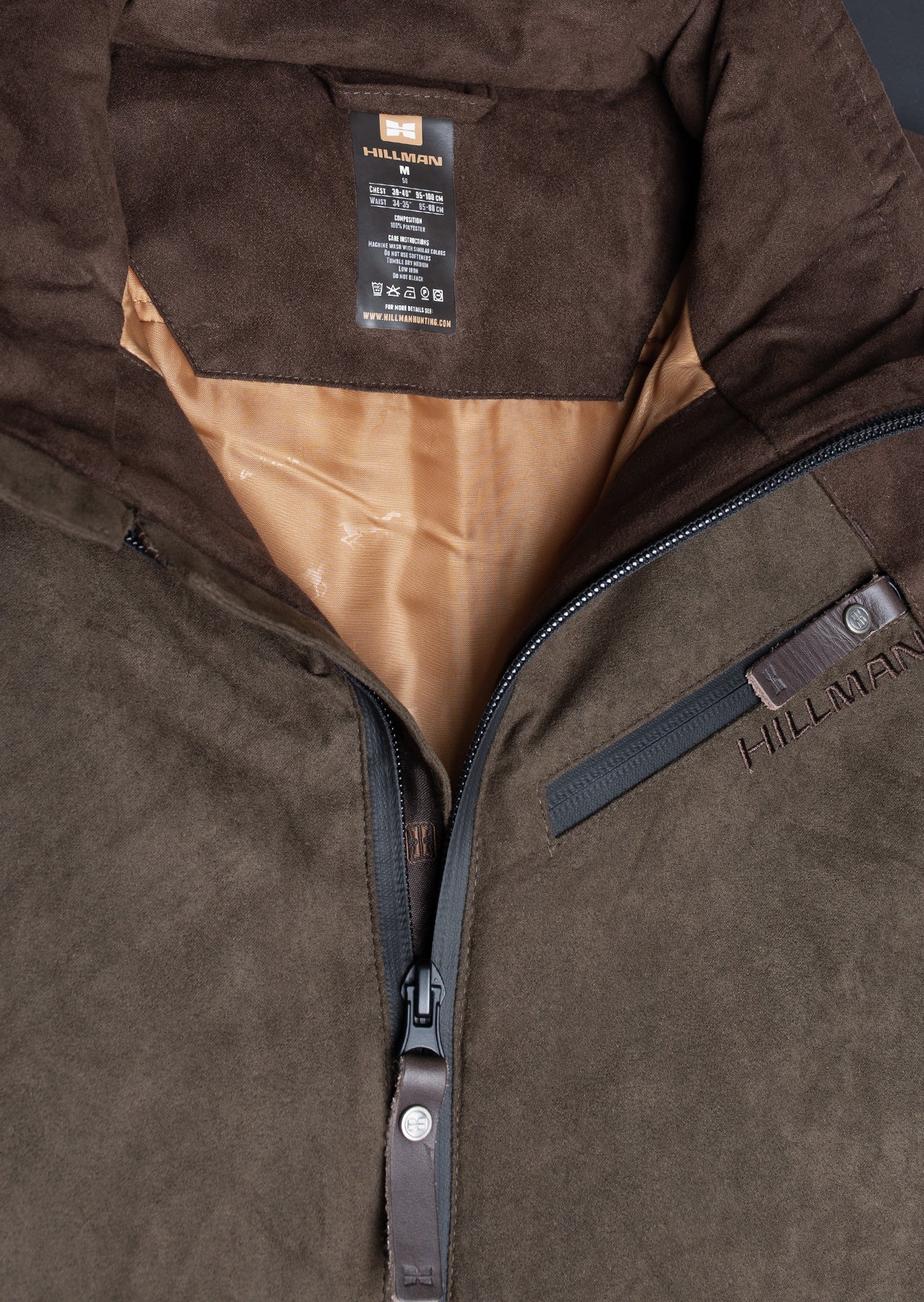 Nahaufnahme der Hillman Fusion™ Jagdjacke Herren Winter. Braunes Veloursleder, ein teilweise geöffneter Reißverschluss mit einer Zuglasche aus Leder, hellbraunes Innenfutter und ein schwarzes Hillman-Etikett heben diese modische wasserdichte und warme Jacke hervor.