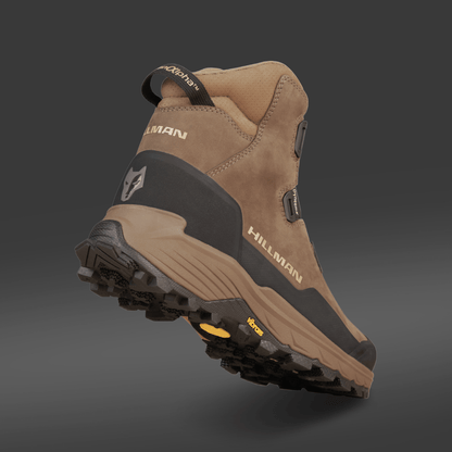 Ein brauner und schwarzer Jagdstiefel Wasserdicht - Herren Jagdschuhe | Alpha DryHunt™ Wanderstiefel von Hillman wird aus einem niedrigen Winkel von hinten auf einem grauen Hintergrund gezeigt, mit einer robusten Sohle und einem Wolfslogo an der Ferse.