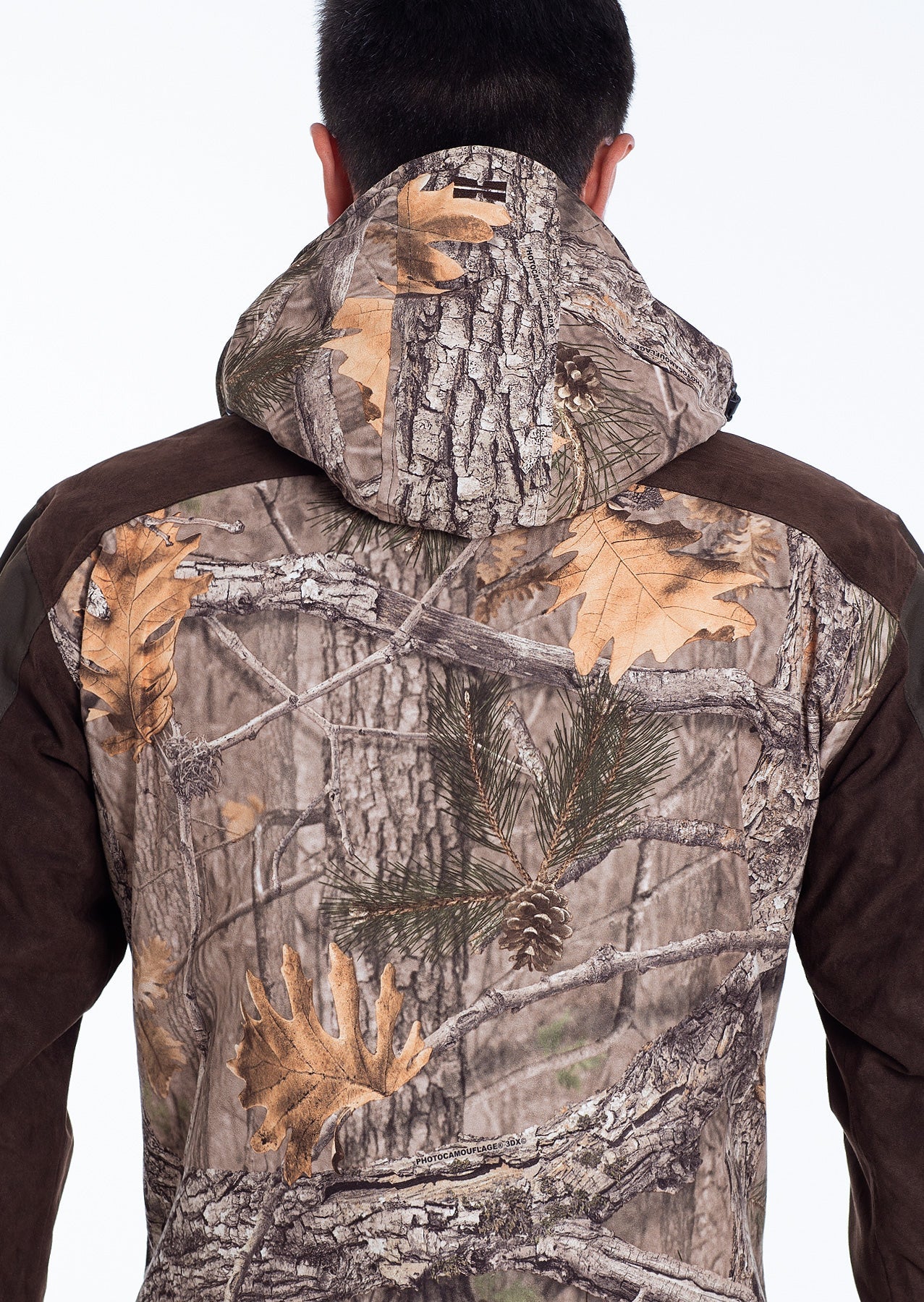 Eine Person, die von hinten gesehen wird, trägt die Hillman Tarnjacke Jagd | Jagdjacke Camouflage - Fusion 3DX mit einer Kapuze, die realistische Baumrinden-, Blatt- und Kiefernnadeldrucke aufweist. Der Hintergrund ist einfarbig weiß.