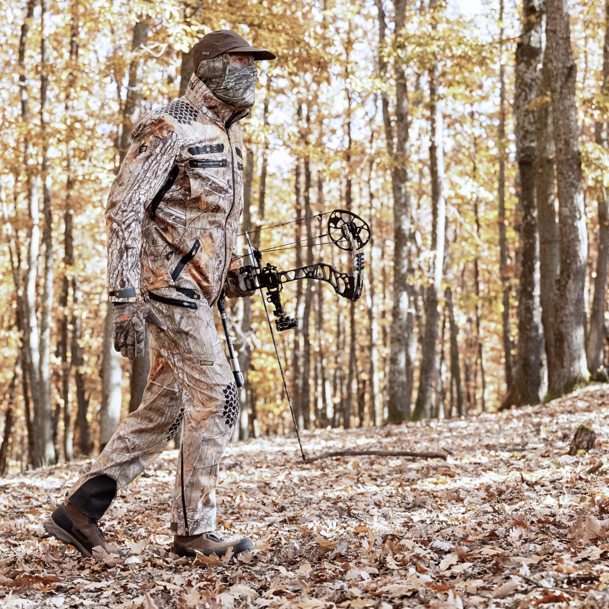 Eine Person in einer Hillman 6OL Winter Jagdhose Camouflage – Tarnhose, wasserdichter Jagdhose und Gesichtsbedeckung geht mit einem Compoundbogen durch einen herbstlichen Wald. Der Boden ist mit Laub bedeckt, umgeben von hohen Bäumen mit spärlichem Laub.