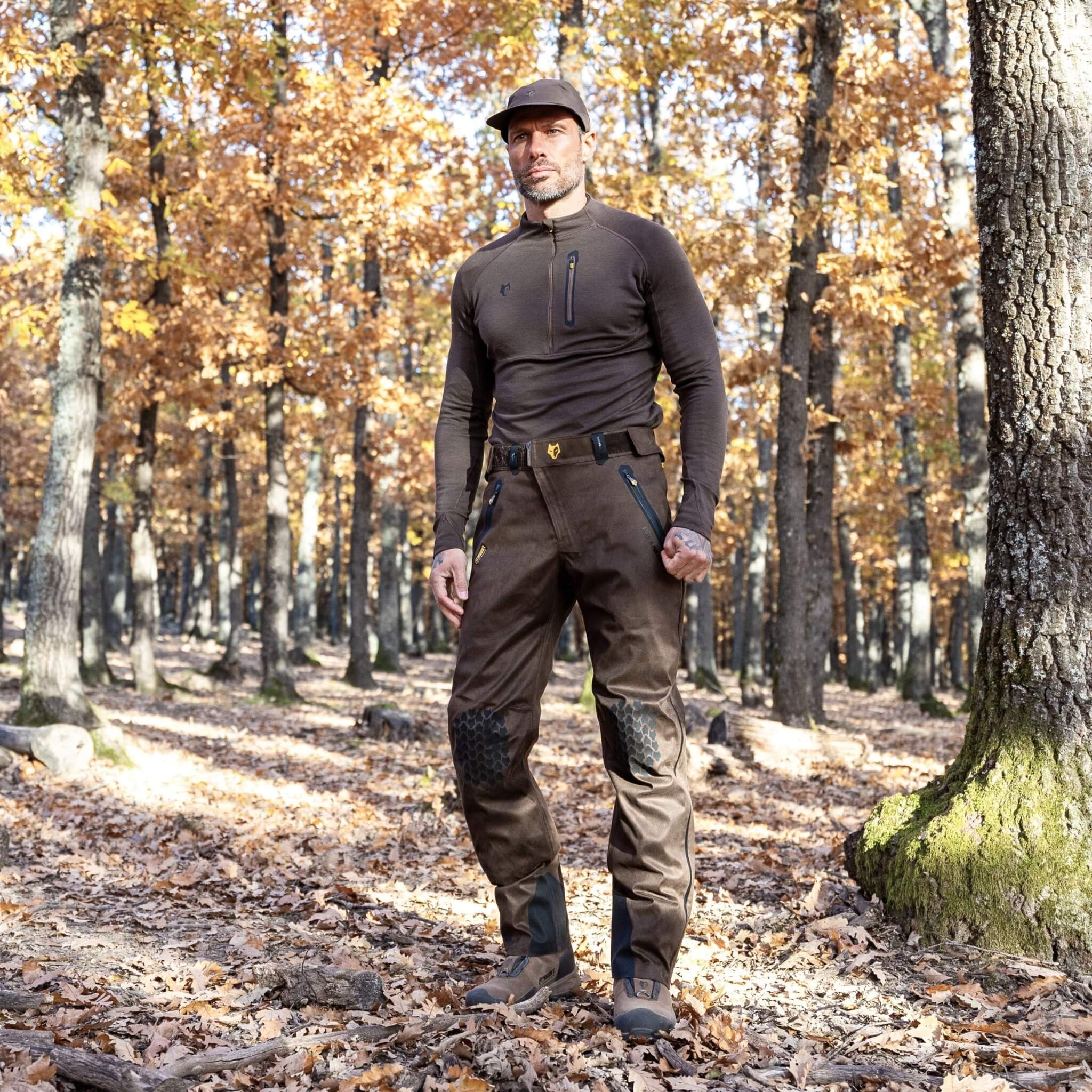 In einem sonnenbeschienenen Wald mit hohen Bäumen und gefallenen Blättern steht eine Person selbstbewusst mit Mütze, Langarmshirt und Hillmans 5WL Wasserdichte Regenhose – Jagdhose Dornenfest und Wasserdicht. Herbstblätter bilden eine warme Kulisse, während Licht durch die Zweige fällt.