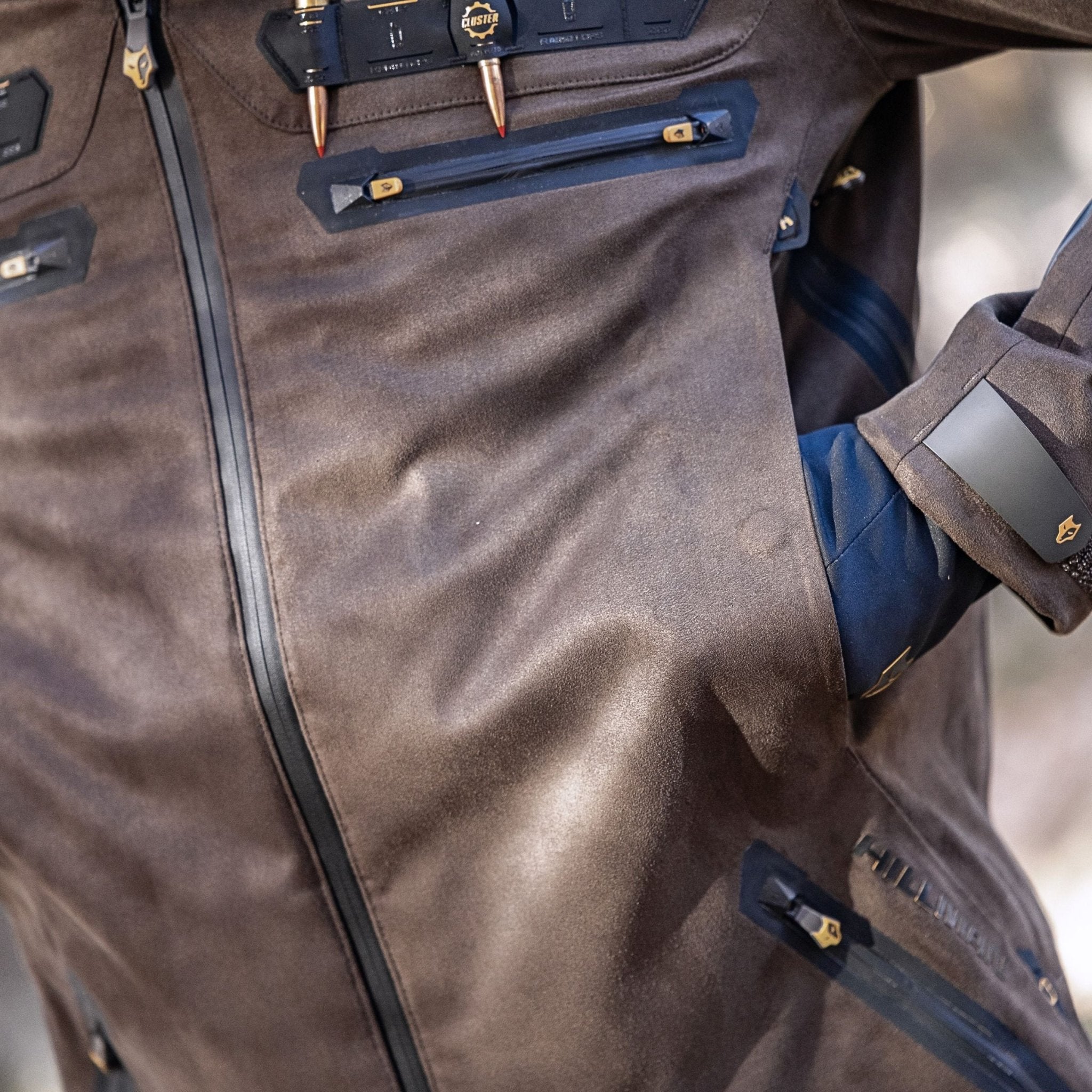 Eine Nahaufnahme zeigt jemanden in einer robusten Hillman 6OL Winter-Jagdjacke in Camouflage – Jagd-Tarnjacke mit schwarzen Reißverschlüssen und mehreren Taschen. Metallische Akzente zieren die Jacke, und in einer Tasche steckt eine Hand in einem blauen Handschuh.