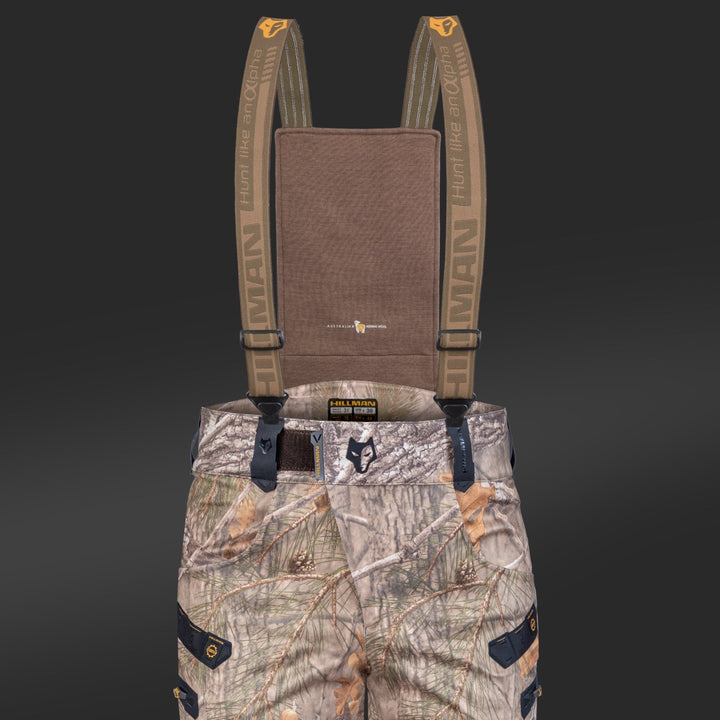 Die Hillman "6OL Winter Jagdhose Camouflage" ist eine wasserdichte Jagdhose mit Hosenträgern im Waldmuster. Die Tarnhose verfügt über mehrere Taschen und eine robuste Textur, ideal für Outdoor-Aktivitäten. Der Hintergrund ist dunkelgrau.