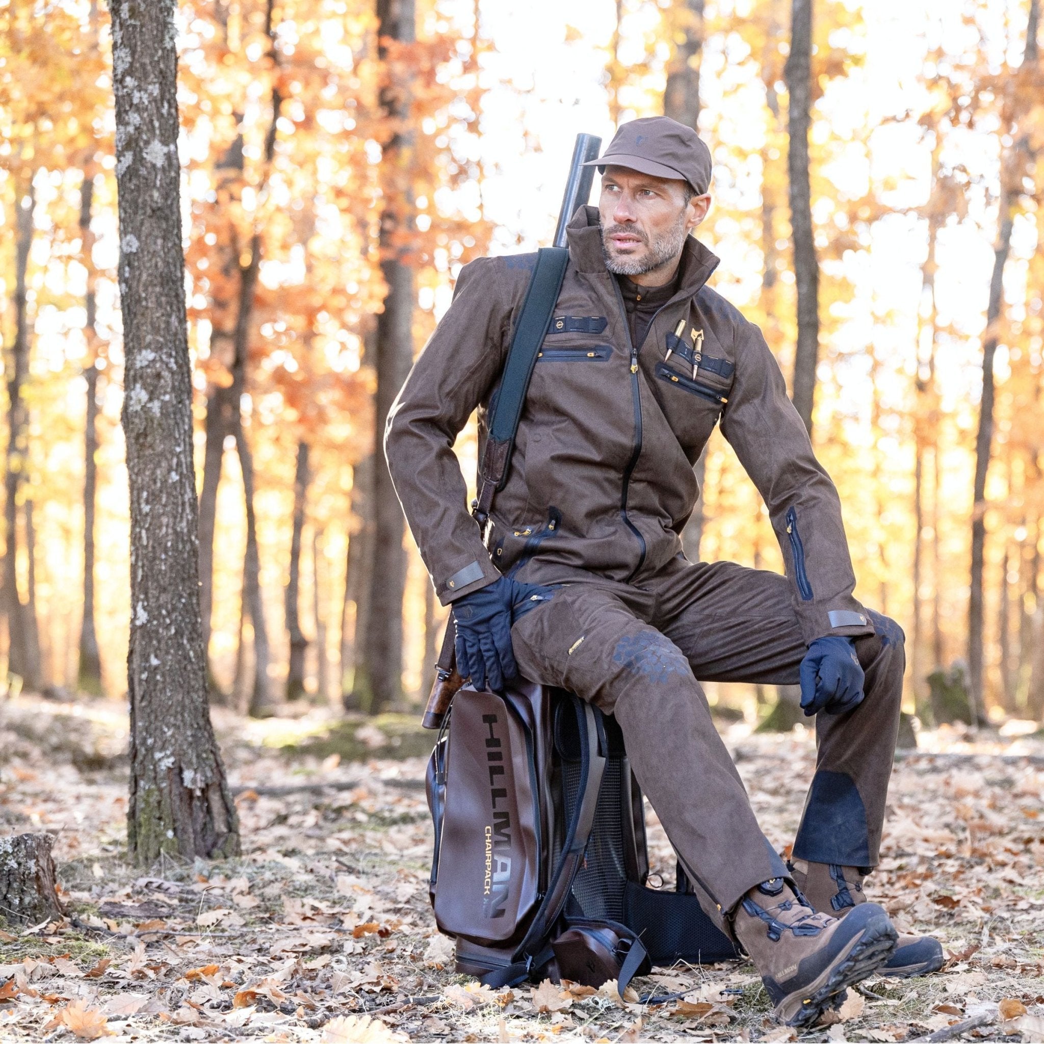 Ein Mann in brauner Jagdkleidung, bekleidet mit der Hillman 6OL Winter Jagdhose Camouflage – Tarnhose, wasserdichter Jagdhose und einer Mütze, sitzt auf einem Baumstumpf im Herbstwald. Er hält ein Gewehr inmitten brauner und orangefarbener Blätter. Ein Rucksack liegt in der Nähe auf dem Boden.