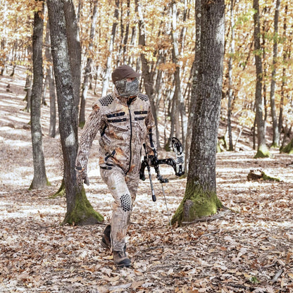Ein Jäger mit Compoundbogen und Hillman 6OL Winterjagdjacke durchquert einen mit Laub bedeckten Wald. Die herbstlichen Bäume bilden eine perfekte Kulisse, sodass sich die Tarnkleidung nahtlos in die Landschaft einfügt.
