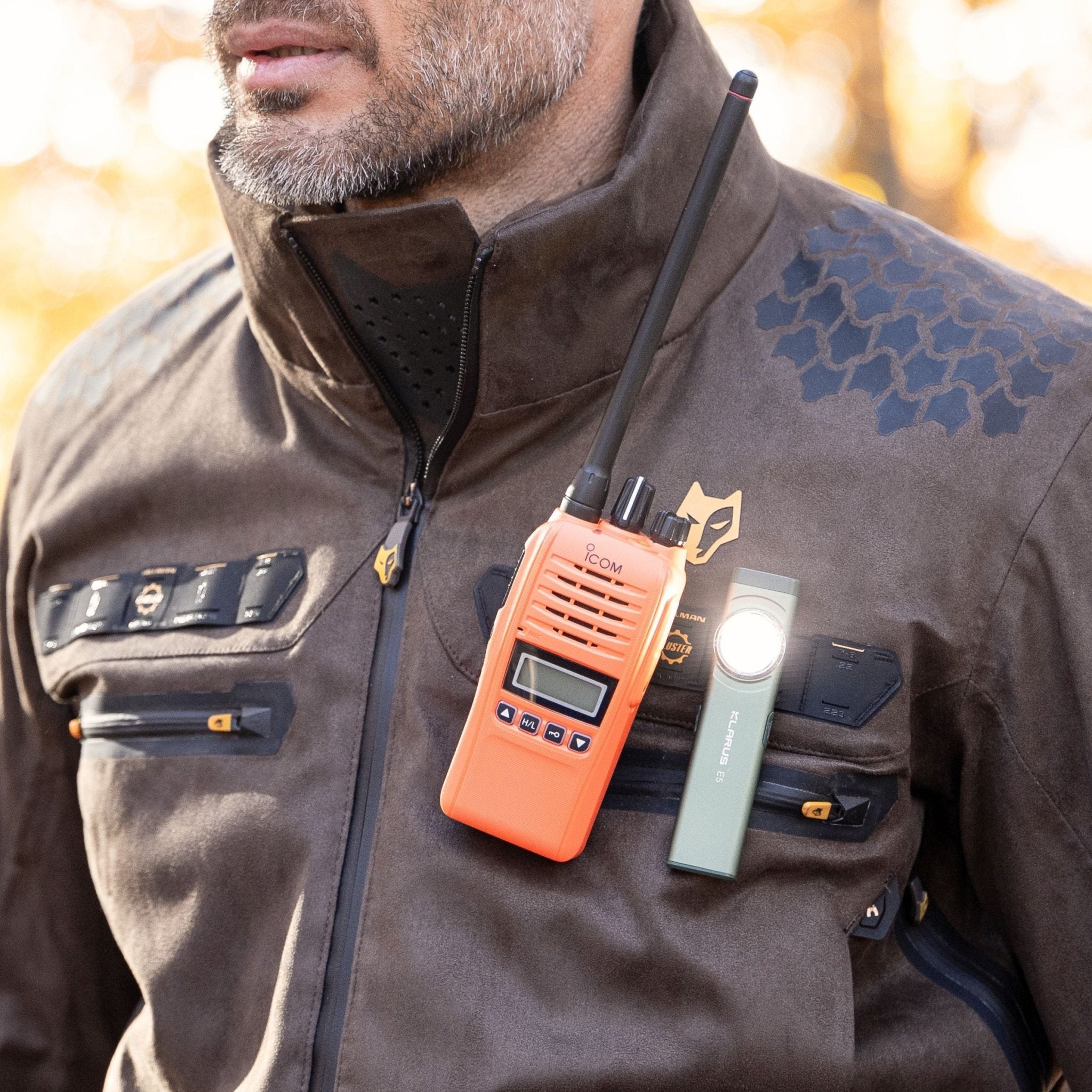 Eine Person in einer Hillman 6OL Winter-Jagdjacke, Tarnmuster mit schwarzen Akzenten, trägt ein leuchtend orangefarbenes Walkie-Talkie und eine kleine Brusttaschenlampe. Die Jacke verfügt über mehrere Reißverschlüsse und ein sechseckiges Muster, das perfekt zur rauen Outdoor-Umgebung passt.