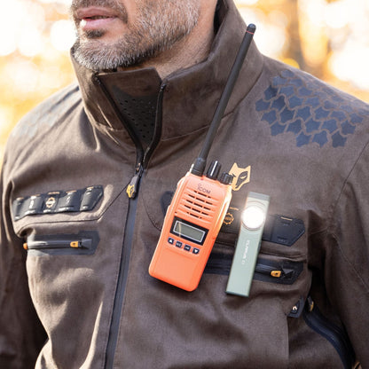 Eine Person in einer Hillman 6OL Winter-Jagdjacke, Tarnmuster mit schwarzen Akzenten, trägt ein leuchtend orangefarbenes Walkie-Talkie und eine kleine Brusttaschenlampe. Die Jacke verfügt über mehrere Reißverschlüsse und ein sechseckiges Muster, das perfekt zur rauen Outdoor-Umgebung passt.