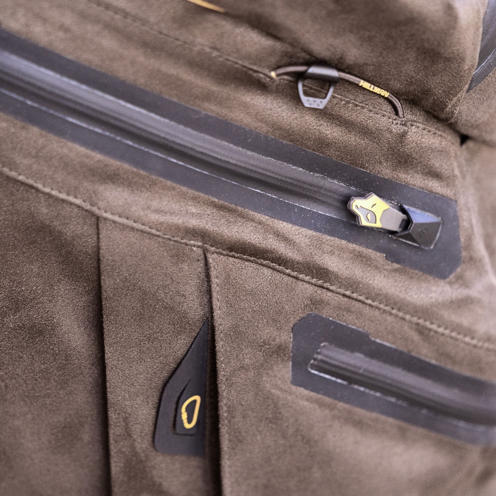 Die Hillman 6OL Winter-Jagdjacke besticht durch eine Nahaufnahme einer braunen Wildlederstruktur mit schwarzen Reißverschlüssen und gelben Akzenten für einen modernen Look. Sichtbare Nähte und kleine lederähnliche Laschen verleihen ihr den letzten Schliff und machen sie perfekt für die Winterjagd.