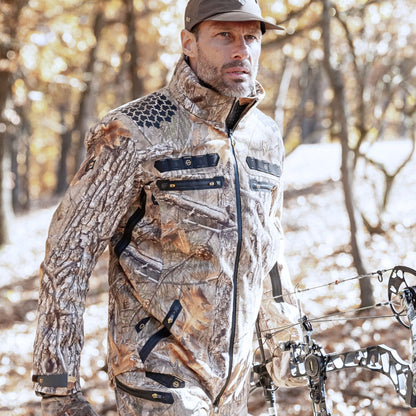 Ein Mann, der eine Hillman 6OL Winter-Jagdjacke – eine wasserdichte Jacke für die Jagd und eine Mütze trägt, trägt einen Compoundbogen, während er durch einen sonnenbeschienenen Wald mit gefallenen Blättern geht.
