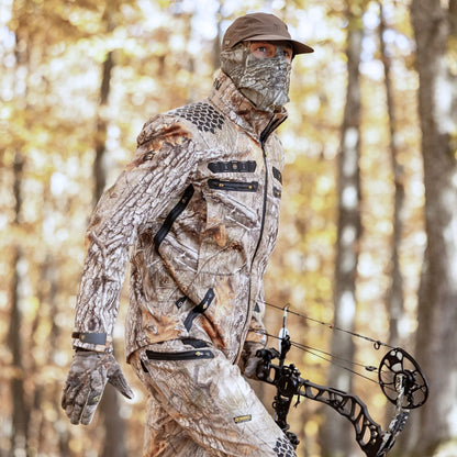 Sie tragen die 6OL Winter-Jagdjacke in Camouflage von Hillman, zusammen mit einer Maske, Handschuhen und einer Mütze, halten einen Compoundbogen und verschmelzen nahtlos mit dem herbstlich gefärbten Wald für ein authentisches Jagderlebnis.
