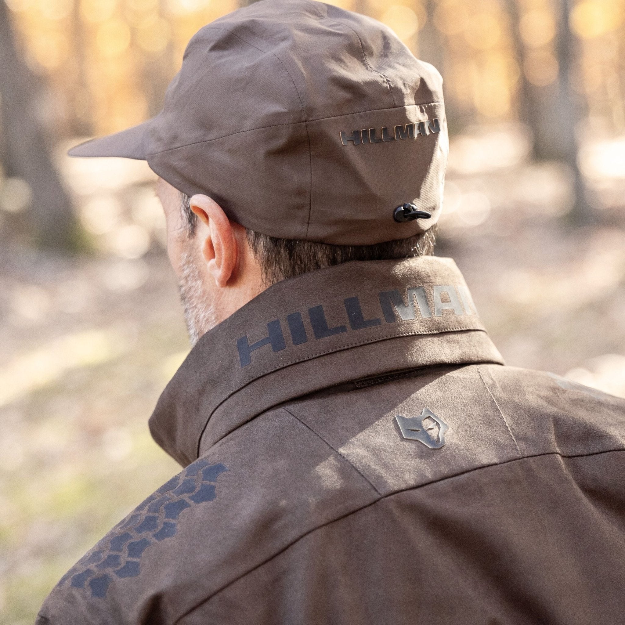 Von hinten ist eine Person zu sehen, die eine braune Mütze und die Hillman 6OL Winterjagdjacke in Camouflage trägt und mit dem herbstlichen Laub eines Waldhintergrunds verschmilzt.