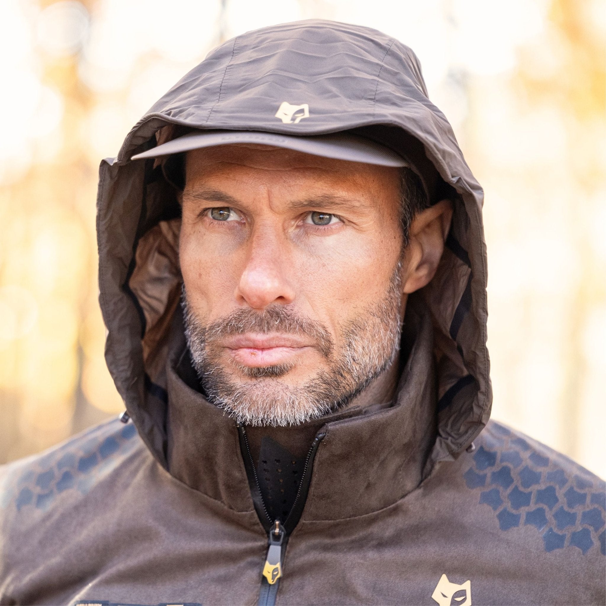 Eine bärtige Person trägt eine Hillman 6OL Winterjagdjacke, einen braunen wasserdichten Jagdmantel mit Kapuze und einen Hut und blickt aufmerksam in die Natur. Der sanft verschwommene Hintergrund ist in warmen Tönen gehalten.