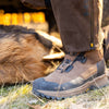 Das Bild zeigt jemanden, der Hillman Vorbestellung 2.0 ALPHA DRYHUNT Jagdstiefel in Braun trägt und auf Gras steht. Der Stiefel ist dafür bekannt, wasserdicht zu sein und drückt sanft gegen das Fell eines Tieres, wobei das Sonnenlicht die Texturen von Leder und Fell hervorhebt.