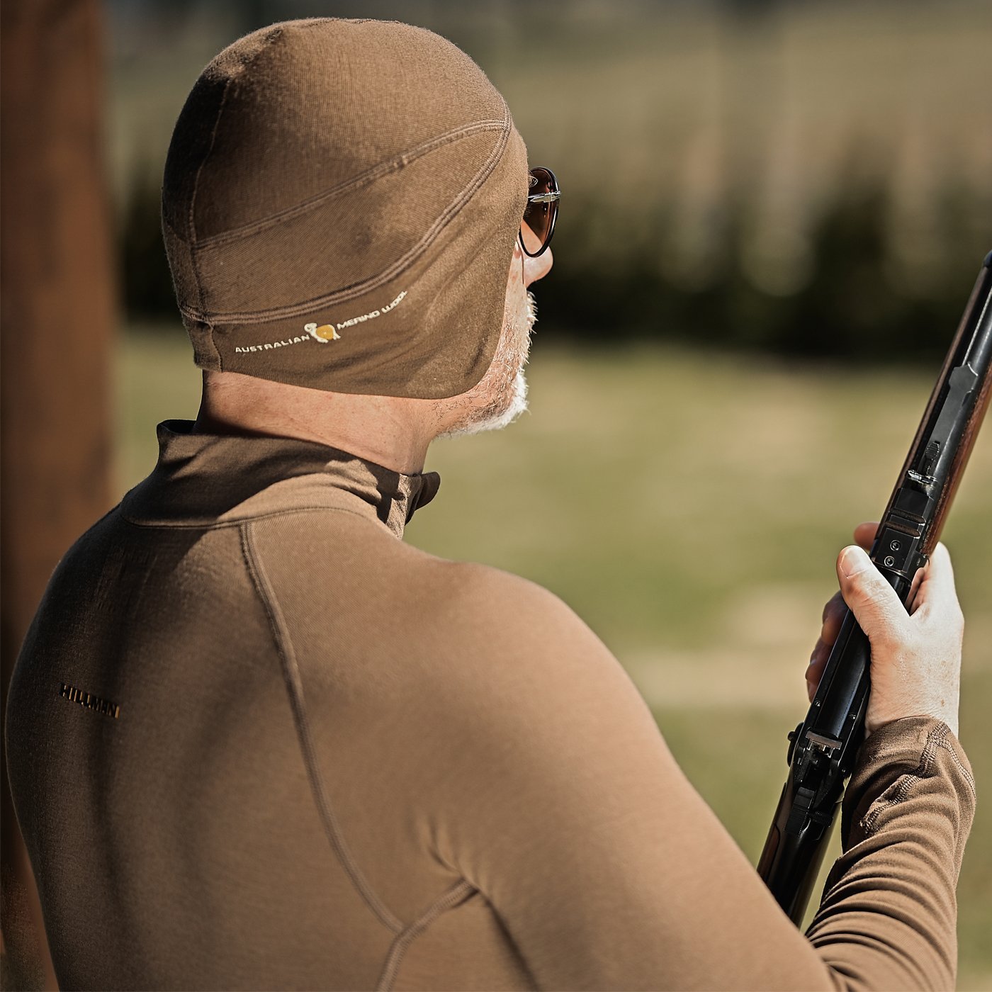 In einer grasbewachsenen Außenumgebung hält eine Person mit einer braunen, angepassten Kappe und einem Oberteil aus Merinowolle zur Wärmeregulierung ein Gewehr. Sie blickt abgewandt, trägt eine Sonnenbrille und die mit dem Logo verzierte [16μm] MERINO THERMOMÜTZE FÜR JAGD von Hillman®GWR. Der unscharfe Hintergrund hebt ihre Präsenz hervor.