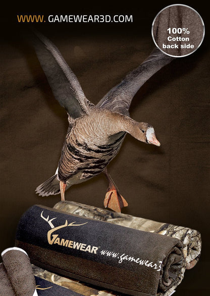 Ein Werbebild zeigt ein Hillman® Duschtuch (70x140cm Blässgans) mit einer Gans, die auf der zusammengerollten Tarndecke landet und das Gamewear-Logo trägt. Oben ist die Website „www.GAMEWEAR3D.com“ zu sehen, und ein Kreis zeigt an, dass die Rückseite der Decke aus 100% Baumwolle besteht.