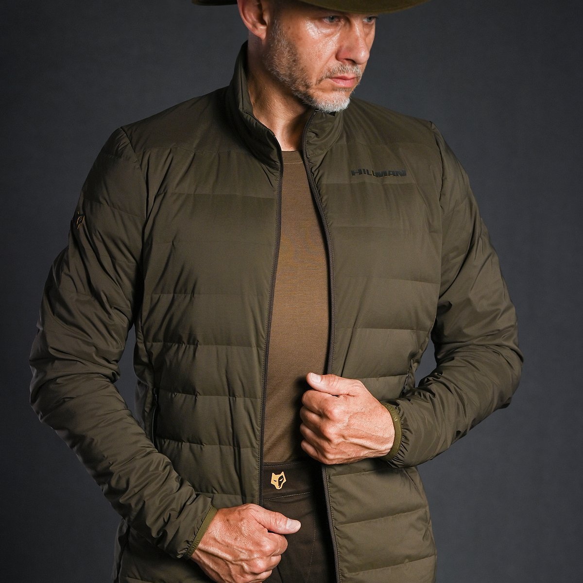Ein Mann in einer braunen Hillman 4IL Winter Daunenjacke mit passender Mütze steht vor einem dunklen Hintergrund. Eine Hand am Reißverschluss, die andere am Saum, rückt er seine für optimale Kälteisolierung konzipierte Jacke zurecht.
